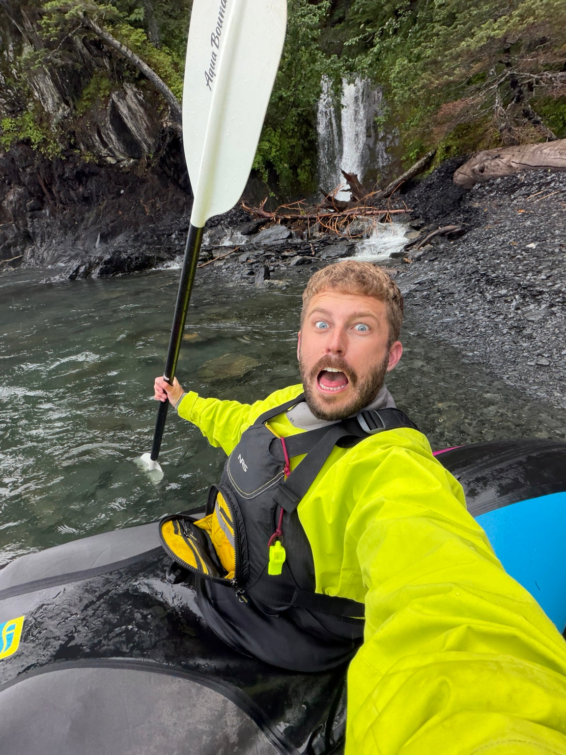 Justin Bickley packrafting beside a waterfall in Alaska wearing a bright yellow paddling top